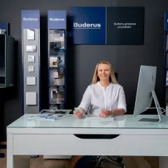 Kobieta przy biurku w salonie Buderus, gdzie oferowany jest serwis pieca gazowego Buderus. W tle ekspozytory z urządzeniami.
