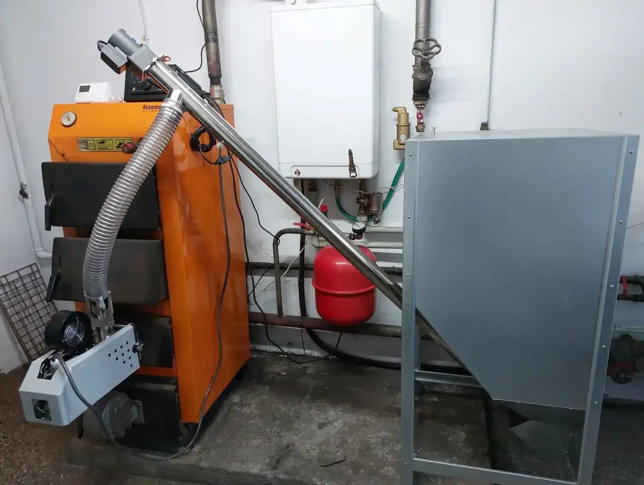 Pomarańczowy piec na ekogroszek z zapalarką, podłączony do zasobnika paliwa i kotła.